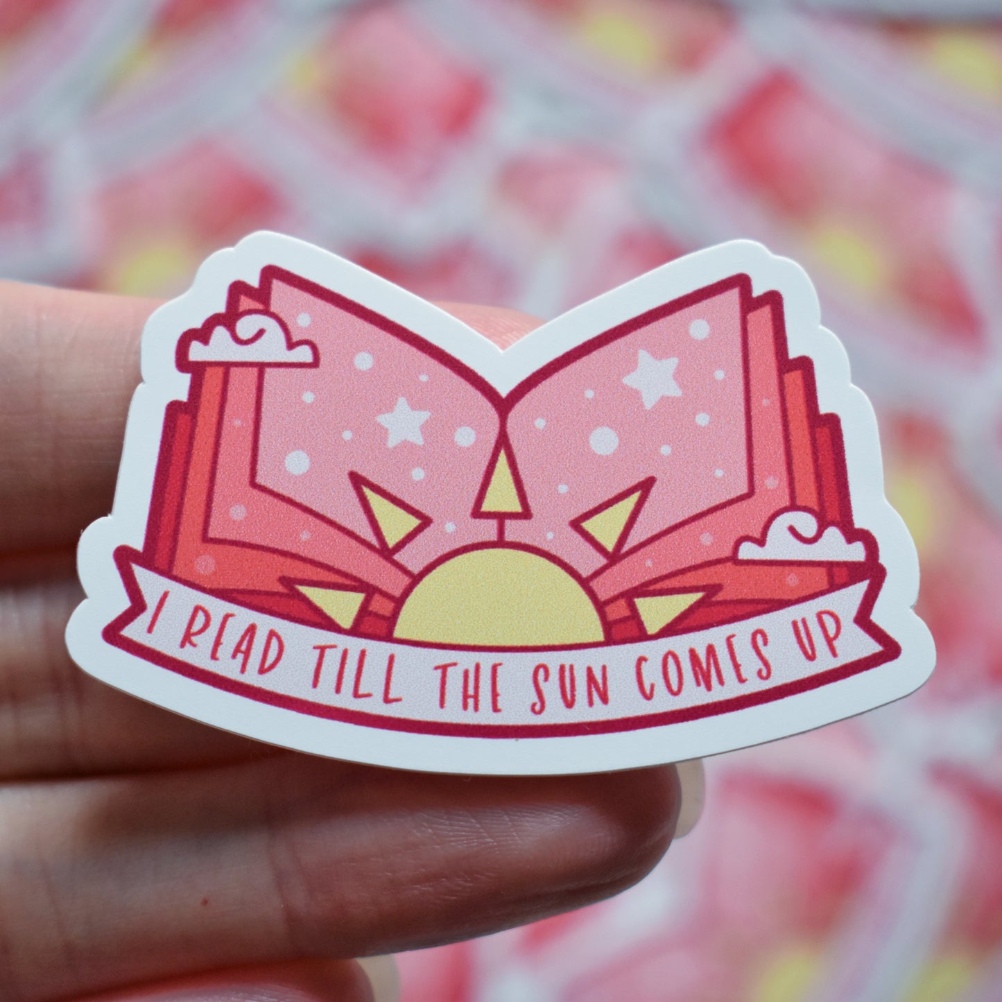 White hand holding sticker. Sticker design is an open book with a pink/peach gradient, with a yellow sun in the middle and white dots/stars/clouds spread across the design. Off white banner at the bottom reads I read till the sun comes up. Sticker has a white border. Background is blurry/distance of same stickers.