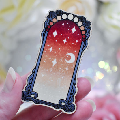 Fingers holding wooden charm without top loop. Design has a black detailed frame. Gradient colour yellow through to red with stars and a moon inside frame.