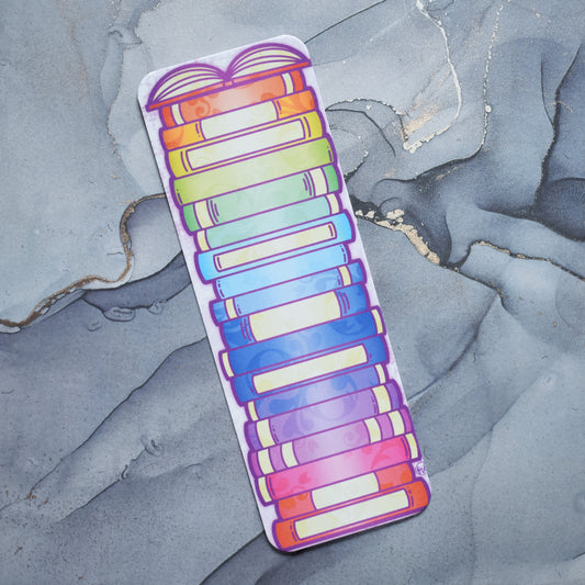 White hand holding bookmark. Bookmark with rounded edges featuring a rainbow bookstack with an open book at the top of the design. Design is slightly off centre on the bookmark. Background is grey watercolour style.