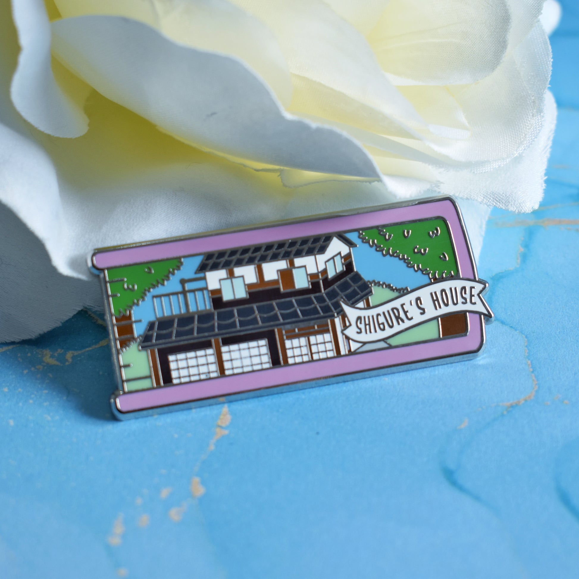 Silver metal enamel pin in centre of image. Enamel pin is shaped like the end of a book in pink and features an old japanese inspired house, in white,brown and black, with traditional sliding doors. Around the house is green grass and big trees. A small banner reads shigure's houseon the bottom right of the design. Background of photograph is blue water inspired with a fake white flower.