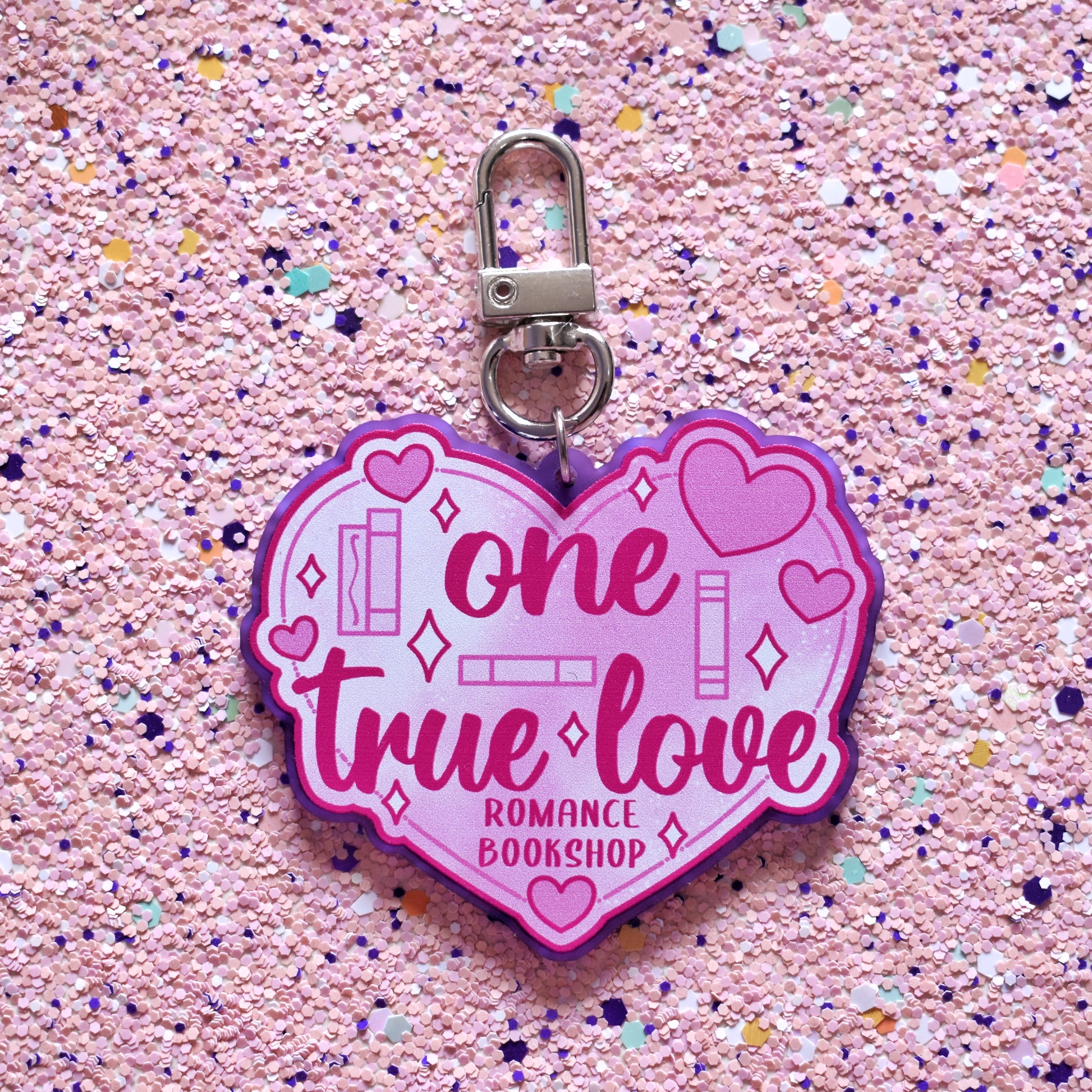 Close up of acrylic keyring in the centre of the image. Featuring a big pink heart with smaller hearts and book spines across the design. Design reads one true love romance bookshop. Keyring is printed on frosted purple acrylic with a silver clasp. The background of the image is peach soft sparkly.