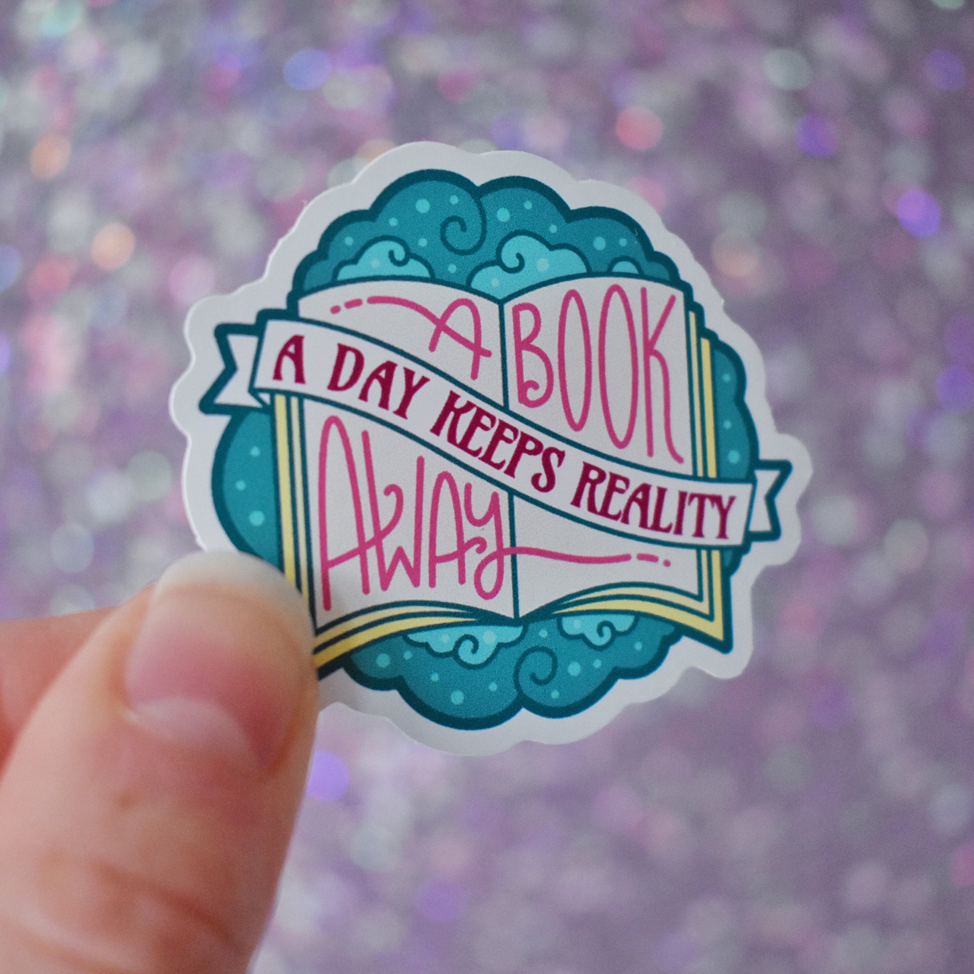 Bookish mini sticker with 'A book a day keeps reality away' text on top of a open book & teal cloud design, tilted at a slight angle, held by a white hand against a glittery purple background