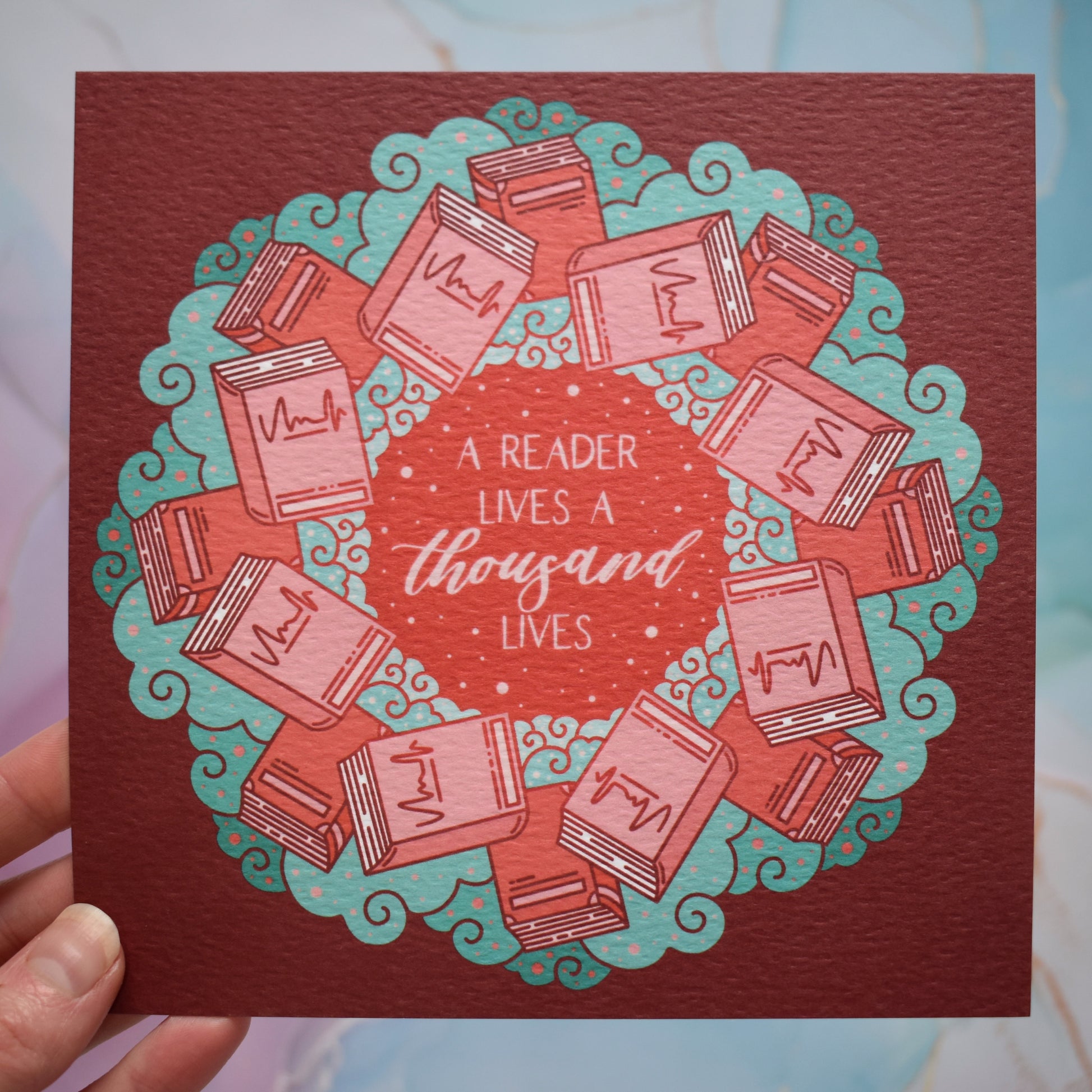White hand holding print. Square print featuring a repeating pattern of red/pink closed books and teal swirls with red around the edge. Design reads a reader lives a thousand lives in pink/red at the centre of the design. Background of photograph is pink/blue watercolour style.