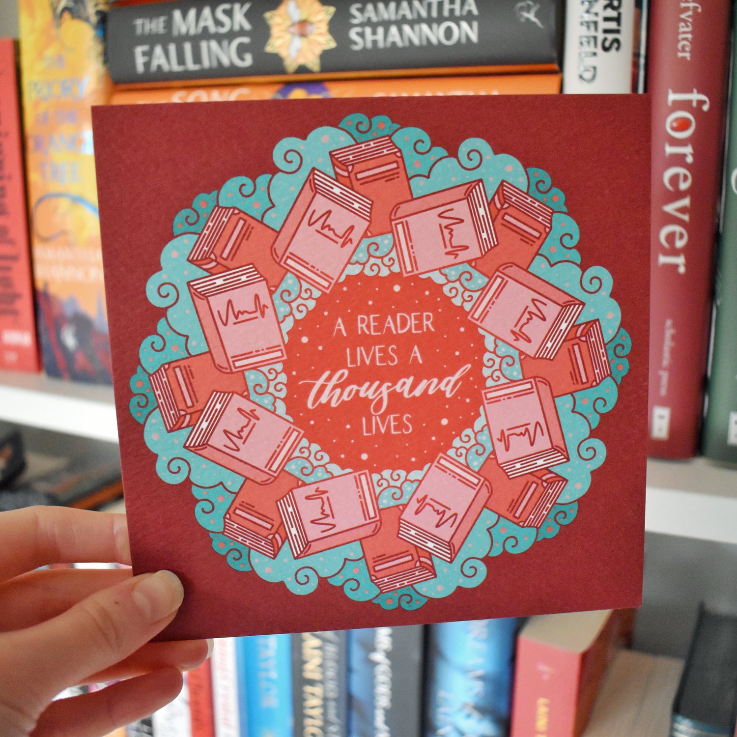 White hand holding print. Square print featuring a repeating pattern of red/pink closed books and teal swirls with red around the edge. Design reads a reader lives a thousand lives in pink/red at the centre of the design. Background of photograph has blurry bookshelf/books.