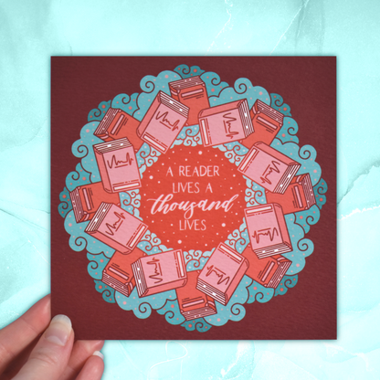 Square bookish print featuring a repeating pattern of red/pink closed books and teal swirls with red around the edge. Design reads 'a reader lives a thousand lives' in pink/red at the centre of the design. Held by a white hand against a background of pastel teal.