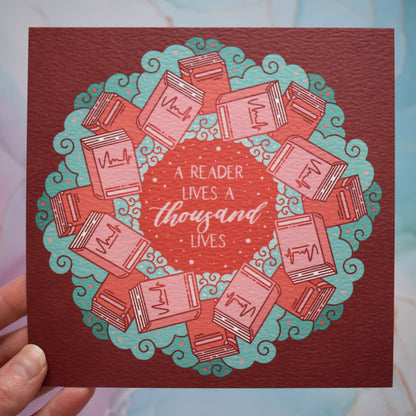 Square bookish print featuring a repeating pattern of red/pink closed books and teal swirls with red around the edge. Design reads 'a reader lives a thousand lives' in pink/red at the centre of the design. Held by a white hand against a background of pink/blue watercolour style.