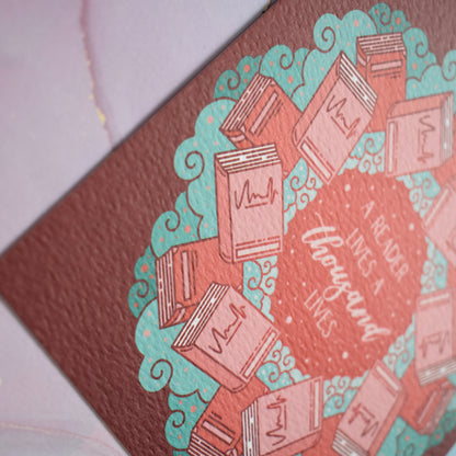Close up of corner of design to show the texture of the print which is slightly raised. Square bookish print featuring a repeating pattern of red/pink closed books and teal swirls with red around the edge. Design reads 'a reader lives a thousand lives' in pink/red at the centre of the design. Background is pink/blue watercolour style.