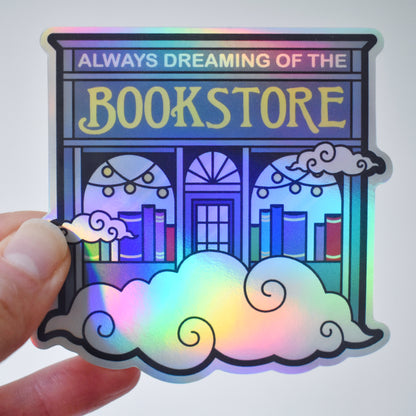 Holographic sticker of a blue bookstore with swirly clouds with 'Always Dreaming of the Bookstore' text. Held by a white hand on an almost white background.