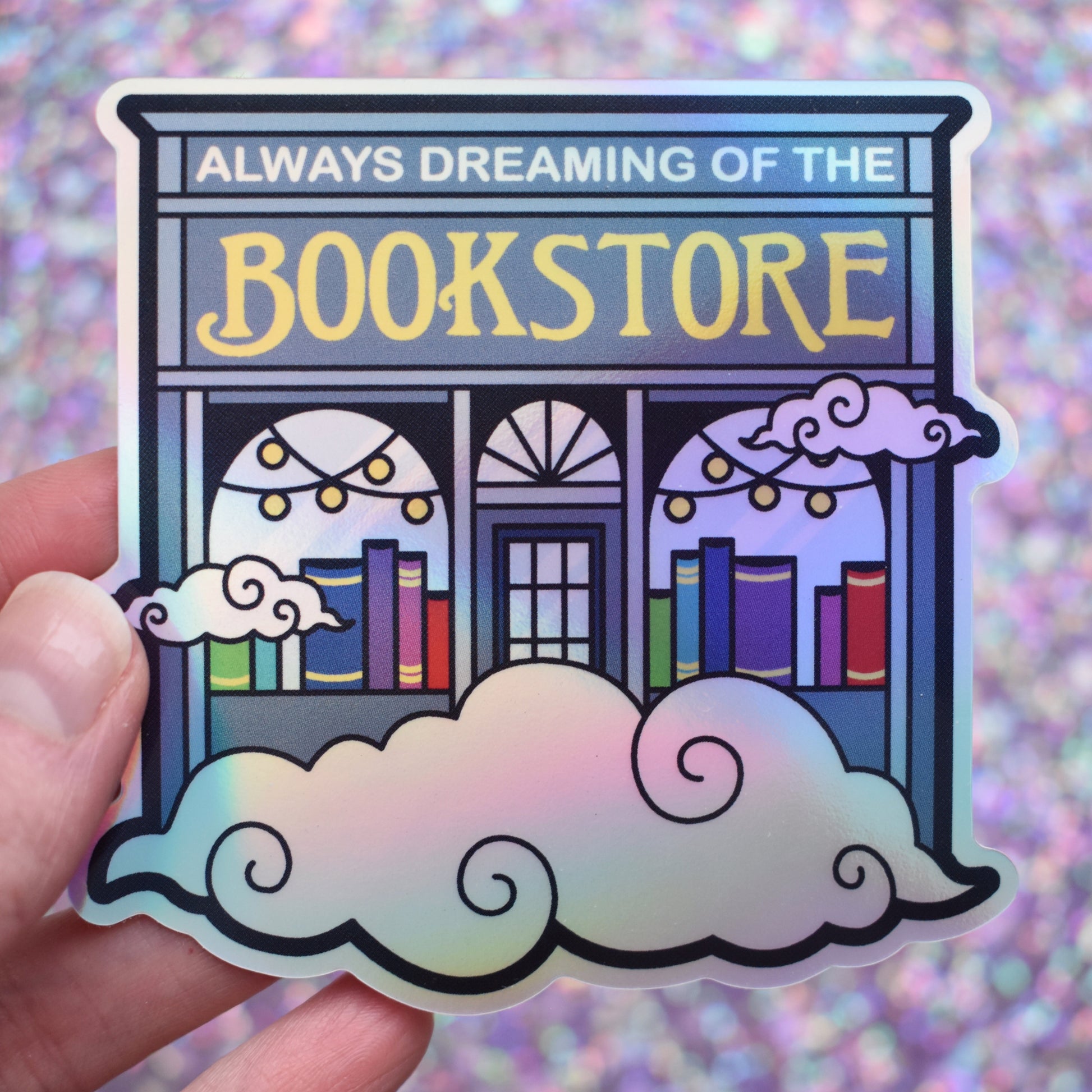 Holographic sticker of a blue bookstore with swirly clouds with 'Always Dreaming of the Bookstore' text held by a white hand on a glittery purple background.