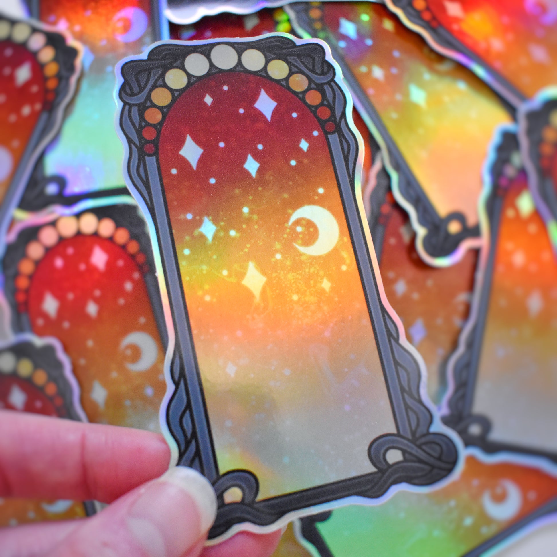 White hand holding sticker tilted to show more holographic effect. Sticker design is a black curly frame with a gradient of circles across the top, from yellowish/peach to red. Centre of the design is a yellowish/peach to red gradient with white/hazy stars and moon. Background is blurry/distance of same stickers.