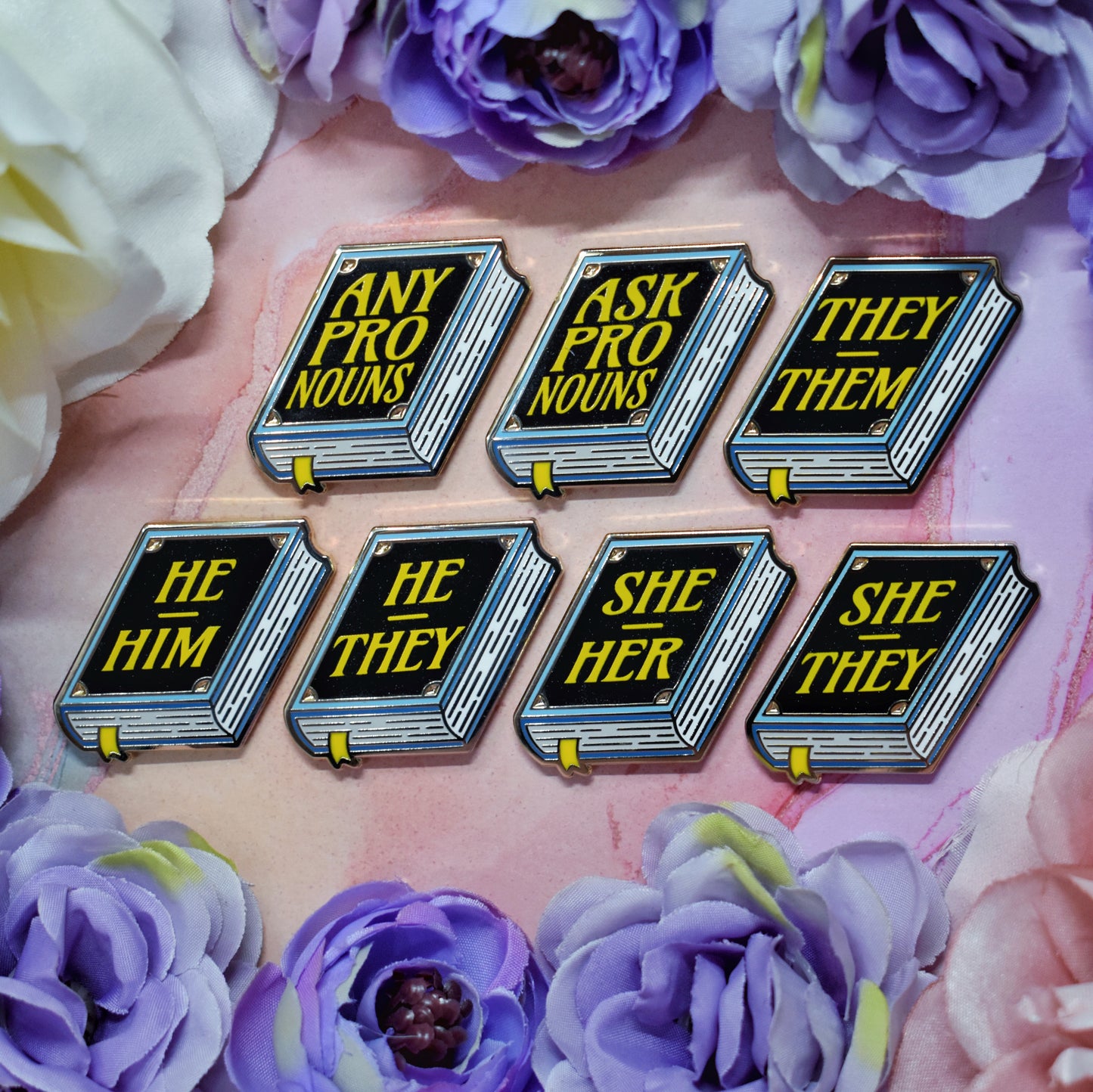 Seven blue, white, black book shaped enamel pins with text 'ANY PRONOUNS' 'ASK PRONOUNS' 'HE HIM', 'HE THEY' 'SHE HER', 'SHE THEY', 'THEY THEM' in blue on a watercolour background with a floral border. 