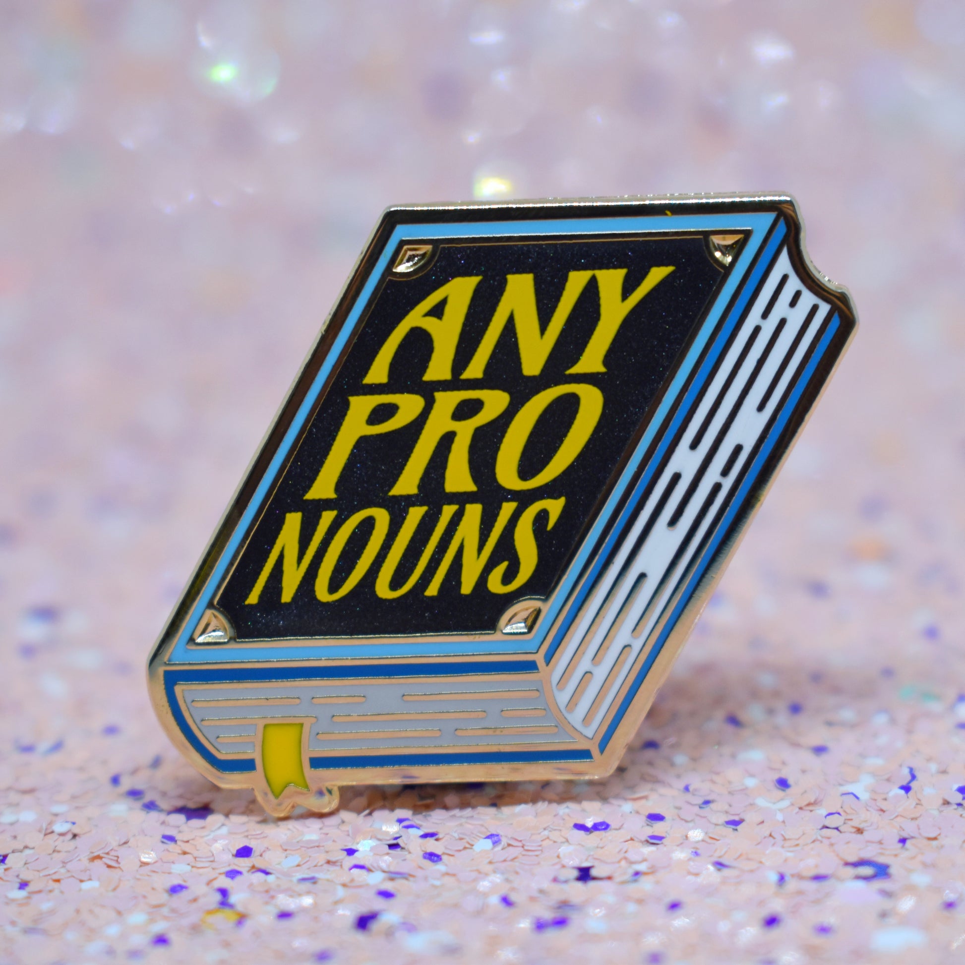 Book shaped enamel pin in gold metal, featuring blue, white and black shimmer accents with 'ANY PRONOUNS' in a bright yellow. Background is a glittery pink with purple specs.