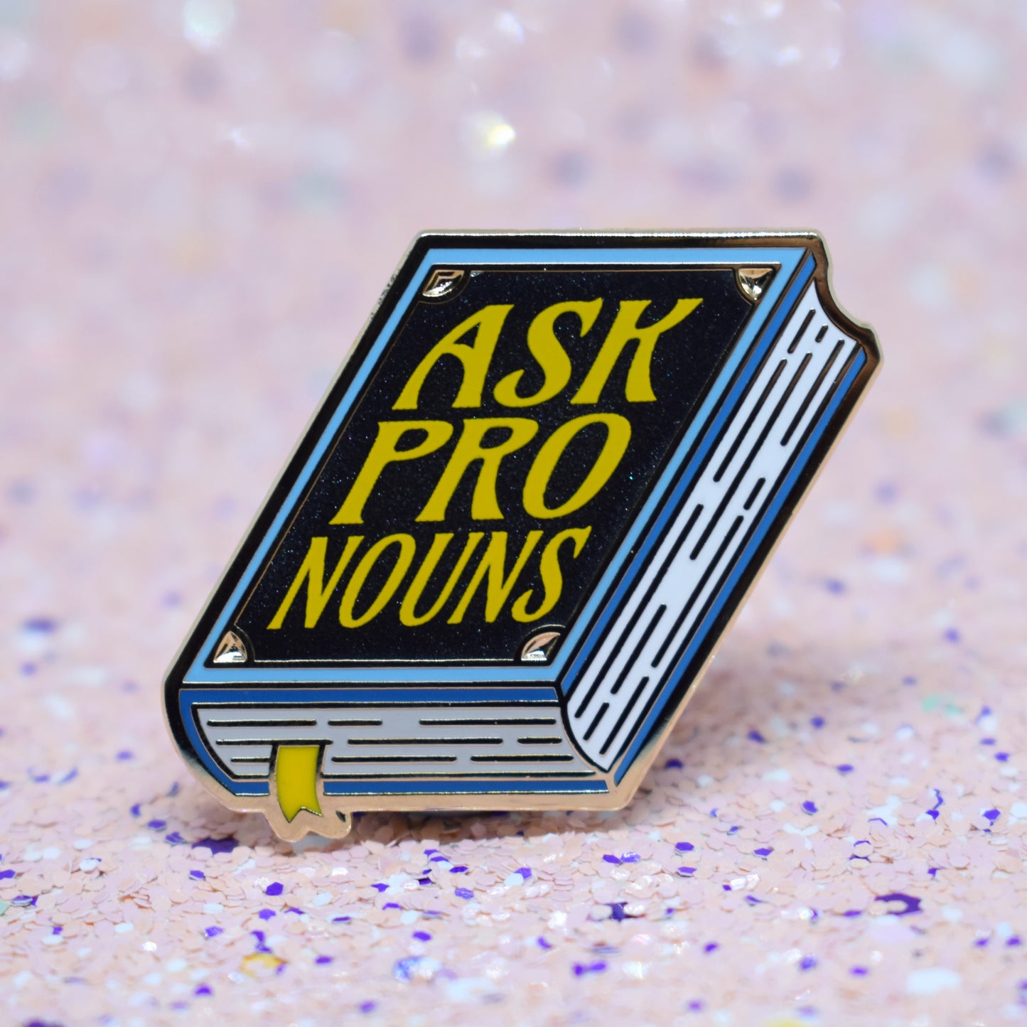 Book shaped enamel pin in gold metal, featuring blue, white and black shimmer accents with 'ASK PRONOUNS' in a bright yellow. Background is a glittery pink with purple specs.