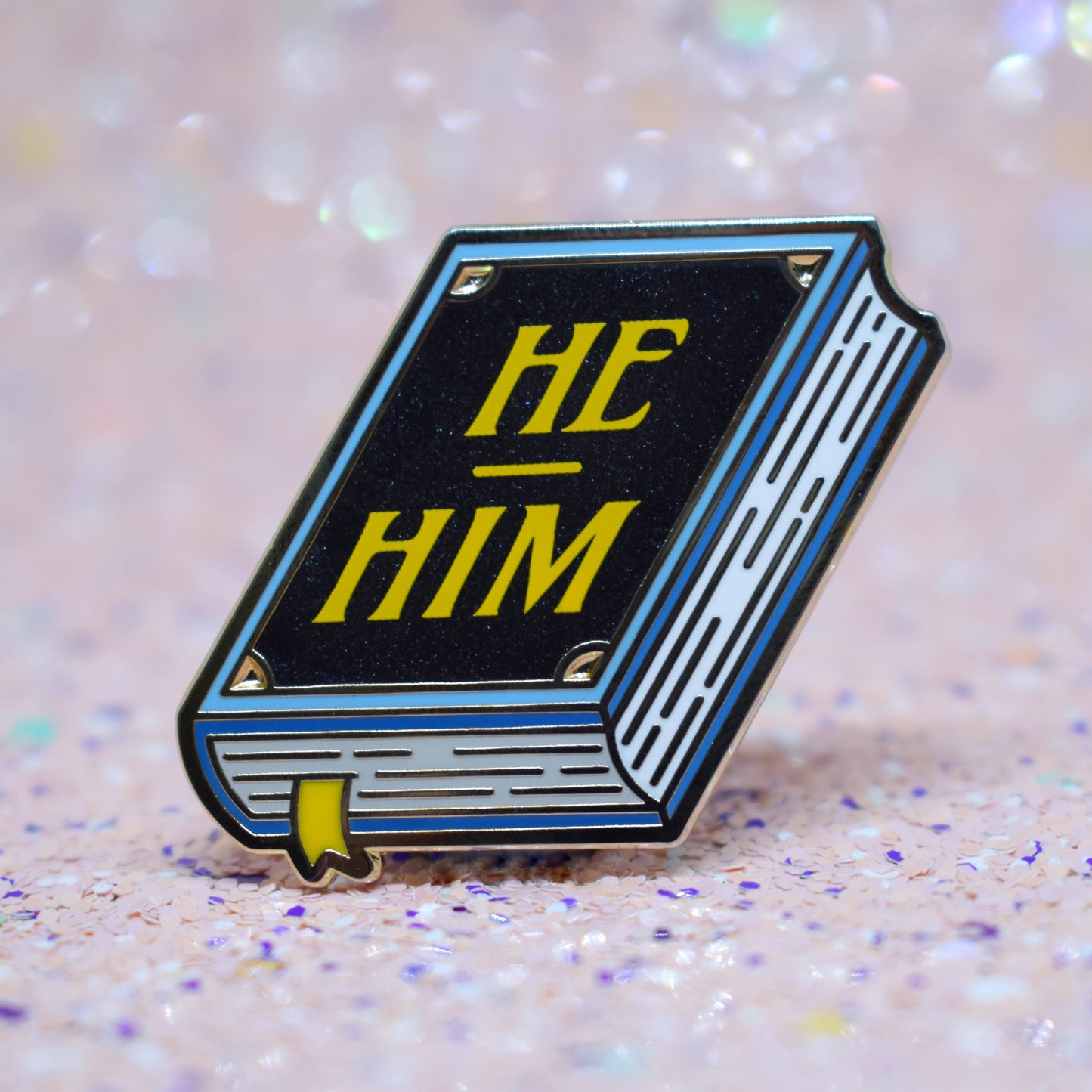 Book shaped enamel pin in gold metal, featuring blue, white and black shimmer accents with 'HE - HIM' in a bright yellow. Background is a glittery pink with purple specs.