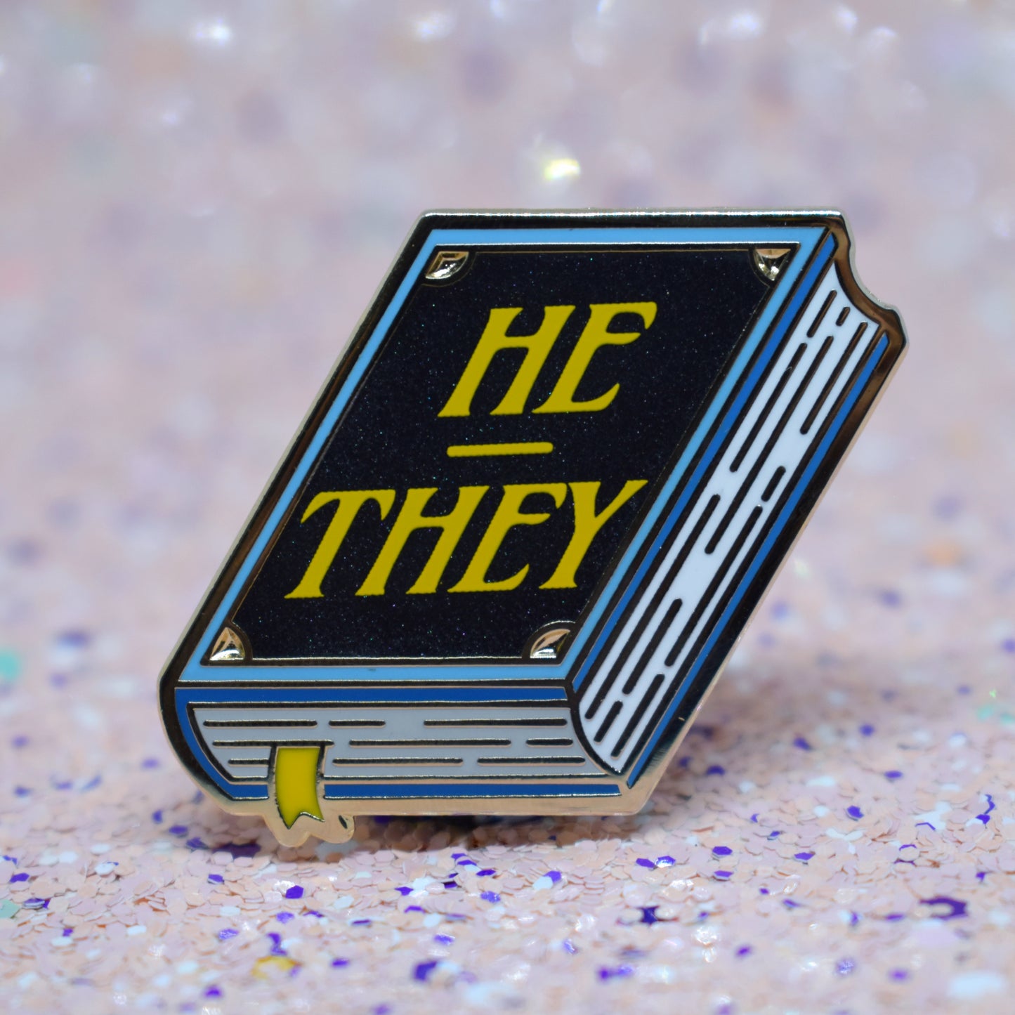 Book shaped enamel pin in gold metal, featuring blue, white and black shimmer accents with 'HE - THEY' in a bright yellow. Background is a glittery pink with purple specs.
