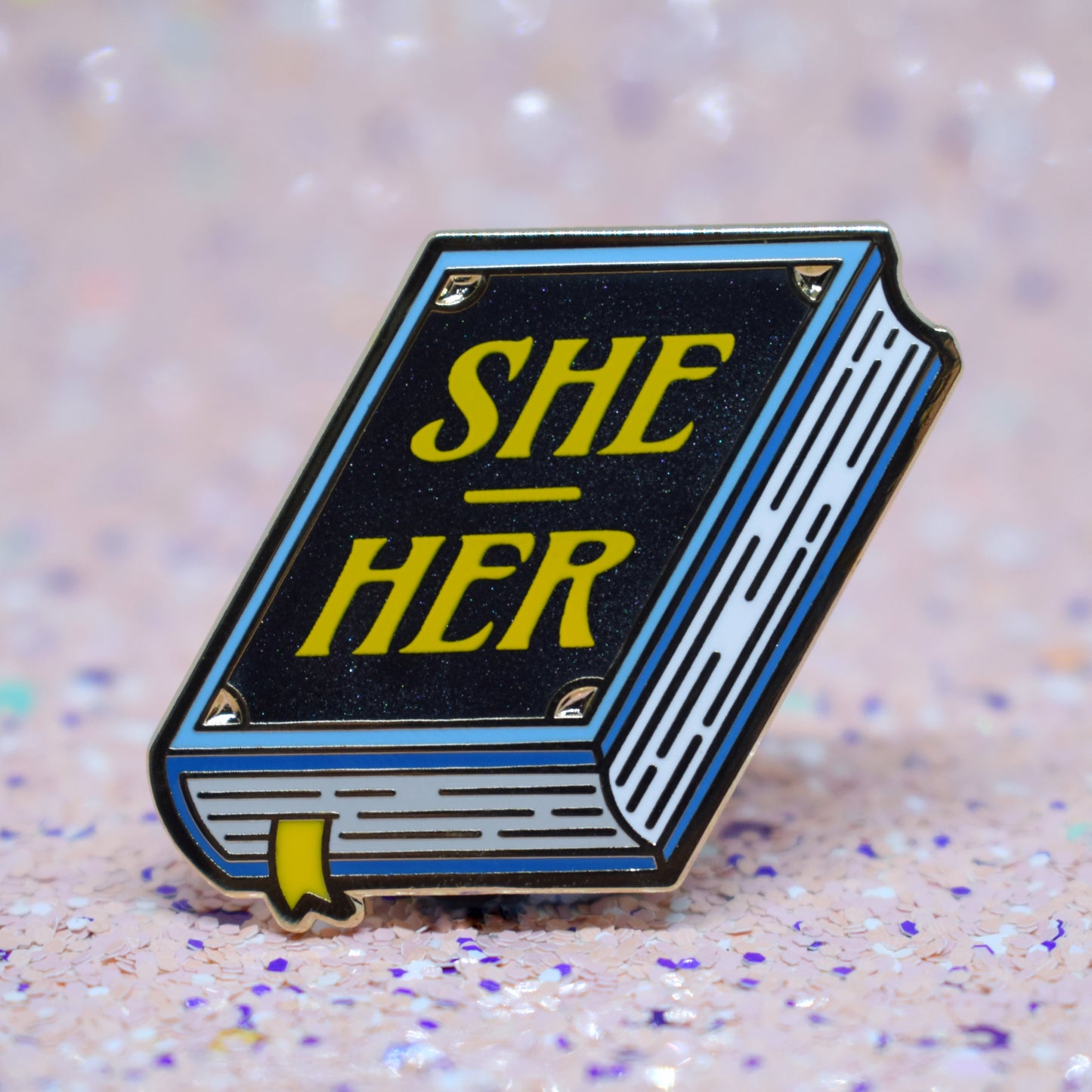 Book shaped enamel pin in gold metal, featuring blue, white and black shimmer accents with 'SHE - HER' in a bright yellow. Background is a glittery pink with purple specs.