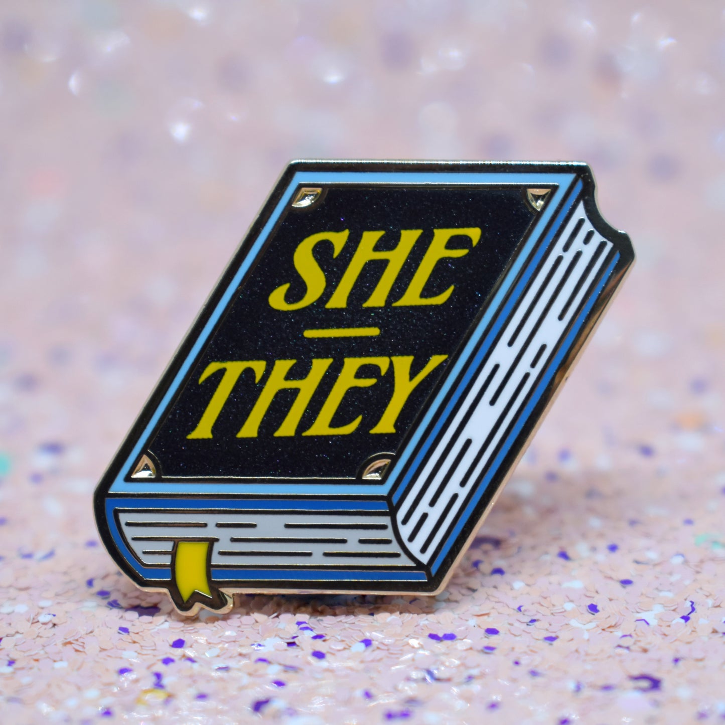 Book shaped enamel pin in gold metal, featuring blue, white and black shimmer accents with 'SHE - THEY' in a bright yellow. Background is a glittery pink with purple specs.