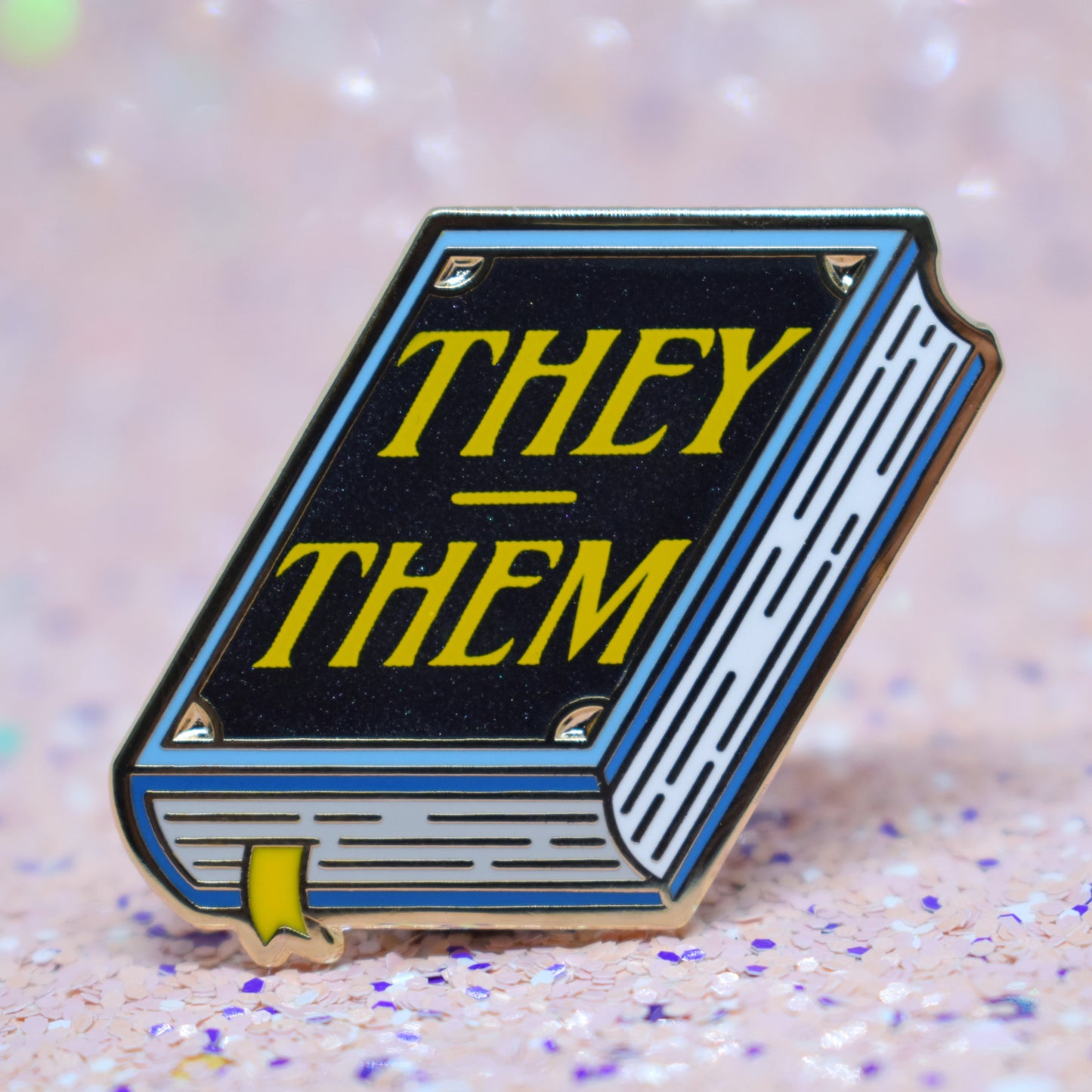 Book shaped enamel pin in gold metal, featuring blue, white and black shimmer accents with 'THEY - THEM' in a bright yellow. Background is a glittery pink with purple specs.