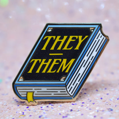 Book shaped enamel pin in gold metal, featuring blue, white and black shimmer accents with 'THEY - THEM' in a bright yellow. Background is a glittery pink with purple specs.
