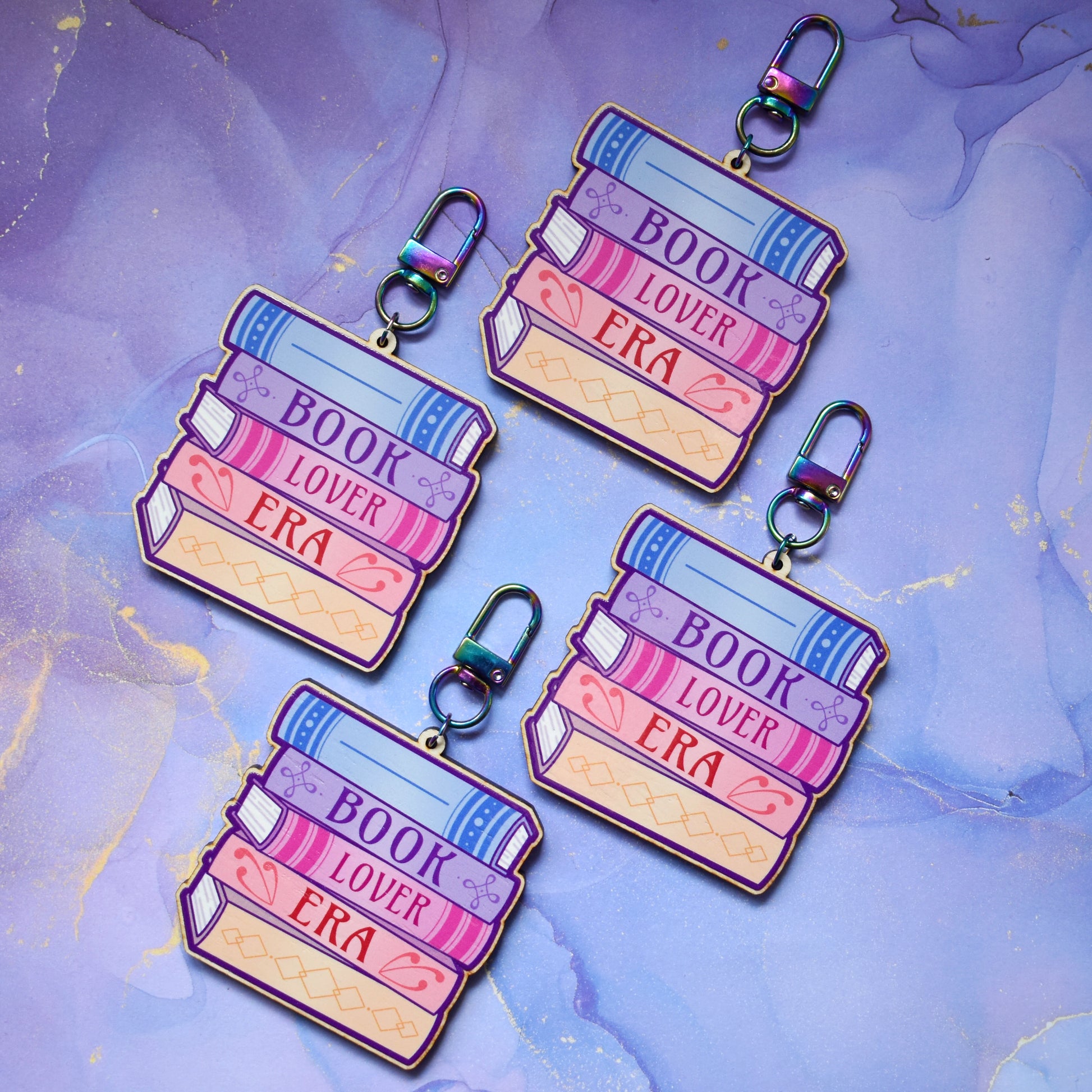 Four wooden keyrings in a slanted diamond shape, with a colourful book stack design with detailed spines and 'Book Lover Era' text. On a colourful watercolour style background.