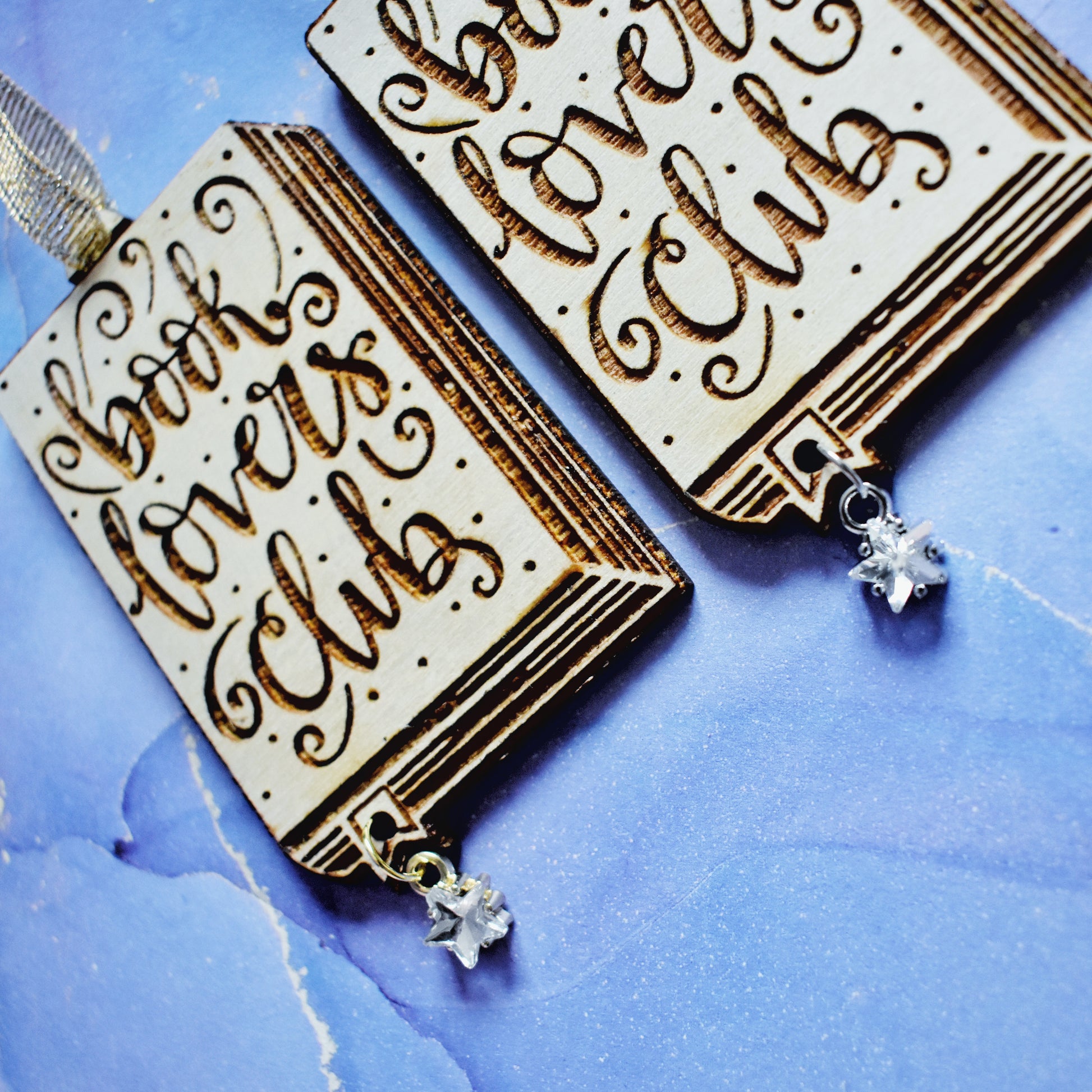 Close up of 2 wooden ornaments next to each other, at an angle on a blue/purple watercolour background. Ornaments are in the shape of a book with book lovers club in loopy writing, showing the page edges at the side and bottom, with a small bookmark end sticking out. A hole in the bookmark end allows for the attachment of a small clear star gem. Left ornamant has a gold ribbon and gold loop on star gem. Right ornamant has a silver ribbon and silver loop on star gem.