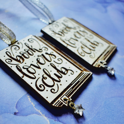 Close up of 2 wooden ornaments next to each other, at an angle on a blue/purple watercolour background. Ornaments are in the shape of a book with book lovers club in loopy writing, showing the page edges at the side and bottom, with a small bookmark end sticking out. A hole in the bookmark end allows for the attachment of a small clear star gem. Left ornamant has a gold ribbon and gold loop on star gem. Right ornamant has a silver ribbon and silver loop on star gem.