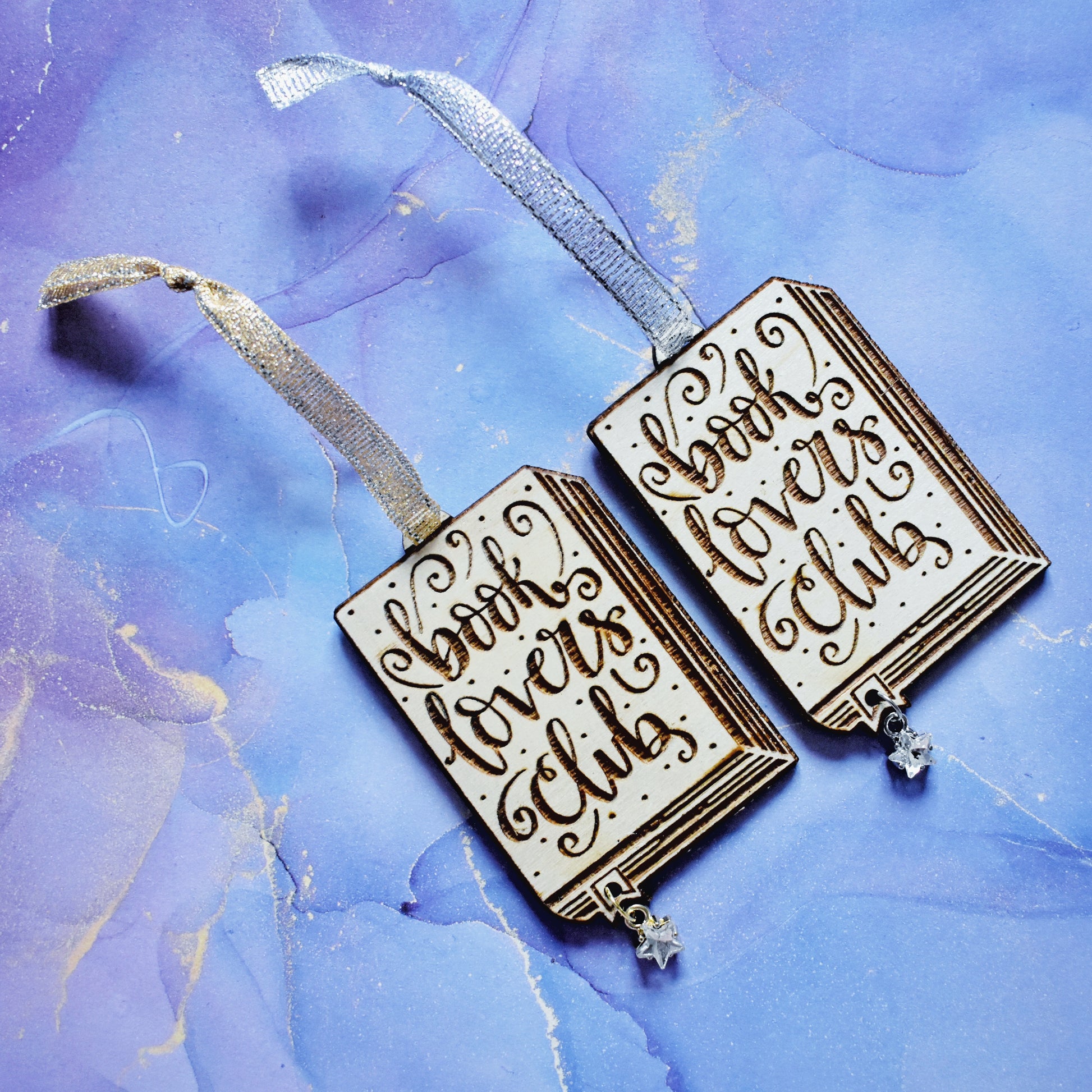 2 wooden ornaments next to each other, at an angle on a blue/purple watercolour background. Ornaments are in the shape of a book with book lovers club in loopy writing, showing the page edges at the side and bottom, with a small bookmark end sticking out. A hole in the bookmark end allows for the attachment of a small clear star gem. Left ornamant has a gold ribbon and gold loop on star gem. Right ornamant has a silver ribbon and silver loop on star gem.
