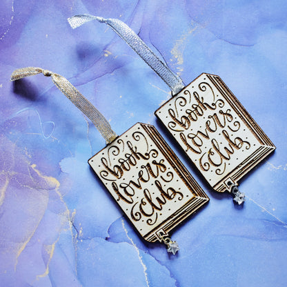 2 wooden ornaments next to each other, at an angle on a blue/purple watercolour background. Ornaments are in the shape of a book with book lovers club in loopy writing, showing the page edges at the side and bottom, with a small bookmark end sticking out. A hole in the bookmark end allows for the attachment of a small clear star gem. Left ornamant has a gold ribbon and gold loop on star gem. Right ornamant has a silver ribbon and silver loop on star gem.