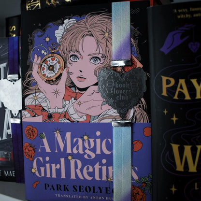 Book titled A Magical Girl Returns is shown with a book band wrapped arpound top to bottom on right hand side. The bookband is pastel rainbow elastic with stainless steel engraved heart and book charm that reads book lovers club. Bookshelf and Other books can be seen in background/forground of image.