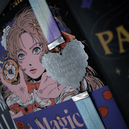 Book titled A Magical Girl Returns is shown with a book band wrapped arpound top to bottom on right hand side. The bookband is pastel rainbow elastic with stainless steel engraved heart and book charm that reads book lovers club. Other books can be seen in background/forground of image.