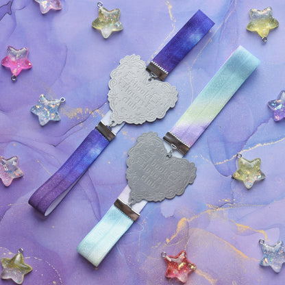 2 book bands diagonal in the centre of image. Top bookband is purple galaxy elastic with stainless steel engraved heart and book charm that reads book lovers club. Bottom bookband is pastel rainbow elastic with stainless steel engraved heart and book charm that reads book lovers club. On a purple/blue watercolour bacground with glittery colourful stars scattered around.
