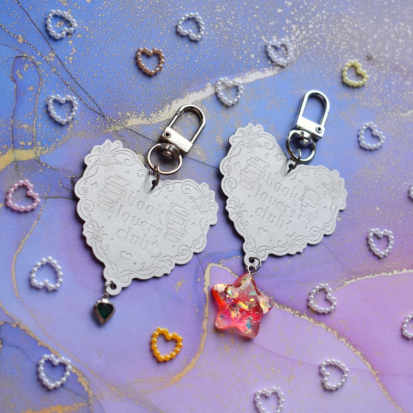 2 keyrings with stainless steel engraved heart and book charm that reads book lovers club at diagonal in the centre of image. Left keyring has a small silver and gem dark green heart attached to charm. Right keyring has a pinky red glittery cabachon star attached to charm. On a pale blue/lilac watercolour background with different coloured heart beads scattered around.