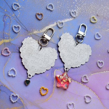 2 keyrings with stainless steel engraved heart and book charm that reads book lovers club at diagonal in the centre of image. Left keyring has a small silver and gem dark green heart attached to charm. Right keyring has a pinky red glittery cabachon star attached to charm. On a pale blue/lilac watercolour background with different coloured heart beads scattered around.