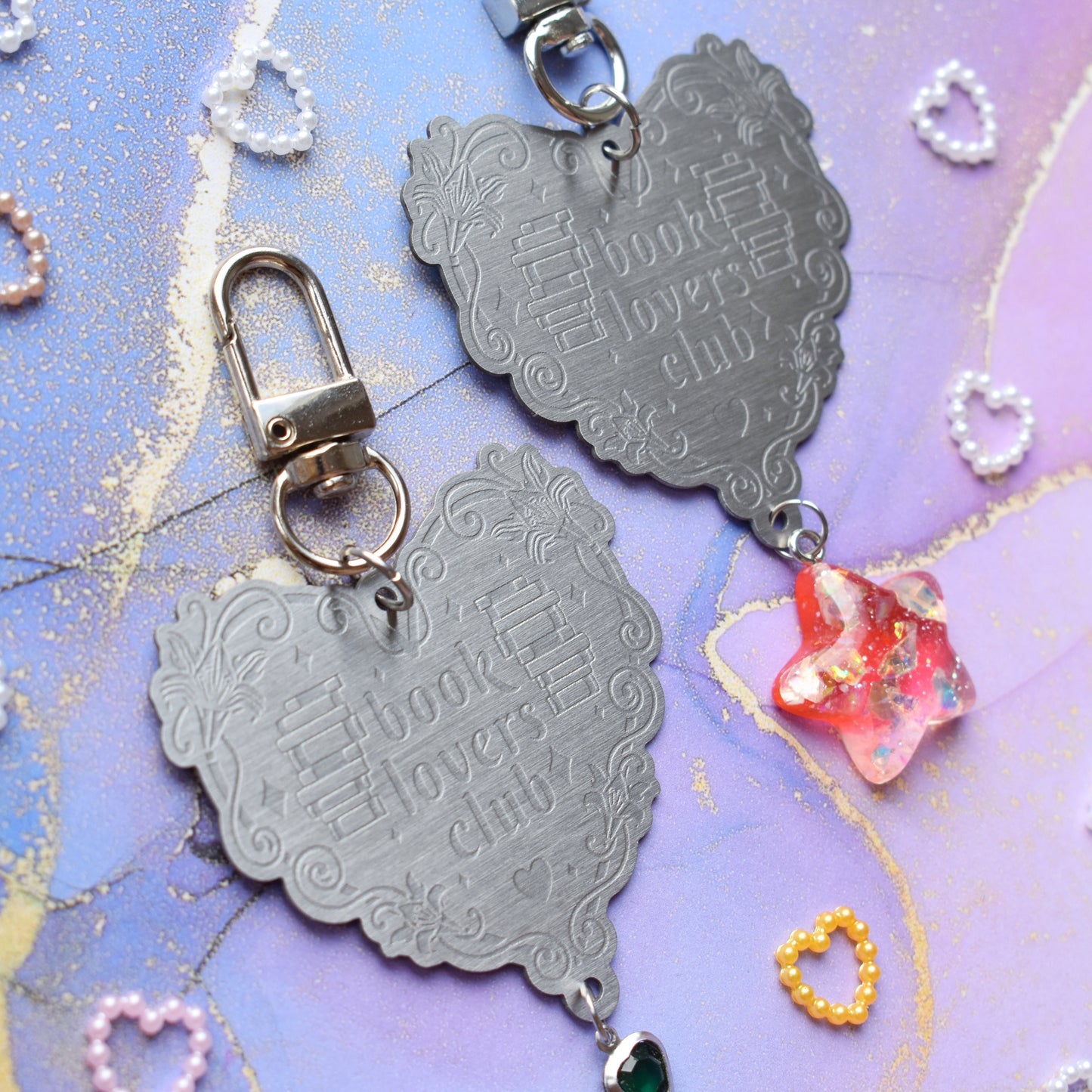 Close up of 2 keyrings with stainless steel engraved heart and book charm that reads book lovers club at diagonal in the centre of image. Left keyring has a small silver and gem dark green heart attached to charm. Right keyring has a pinky red glittery cabachon star attached to charm. On a pale blue/lilac watercolour background with different coloured heart beads scattered around.