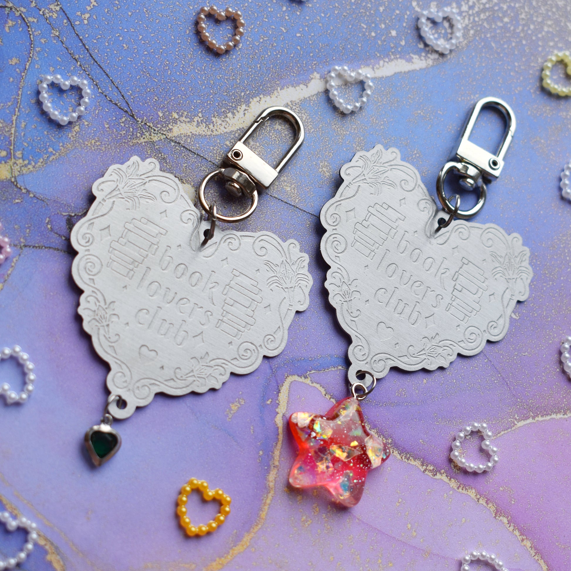 2 keyrings with stainless steel engraved heart and book charm that reads book lovers club at diagonal in the centre of image. Left keyring has a small silver and gem dark green heart attached to charm. Right keyring has a pinky red glittery cabachon star attached to charm. On a pale blue/lilac watercolour background with different coloured heart beads scattered around.