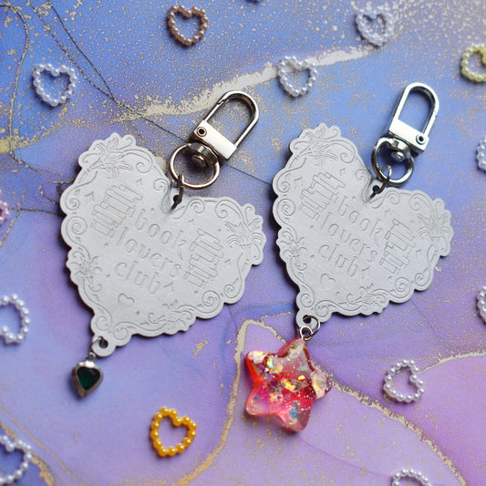 2 keyrings with stainless steel engraved heart and book charm that reads book lovers club at diagonal in the centre of image. Left keyring has a small silver and gem dark green heart attached to charm. Right keyring has a pinky red glittery cabachon star attached to charm. On a pale blue/lilac watercolour background with different coloured heart beads scattered around.