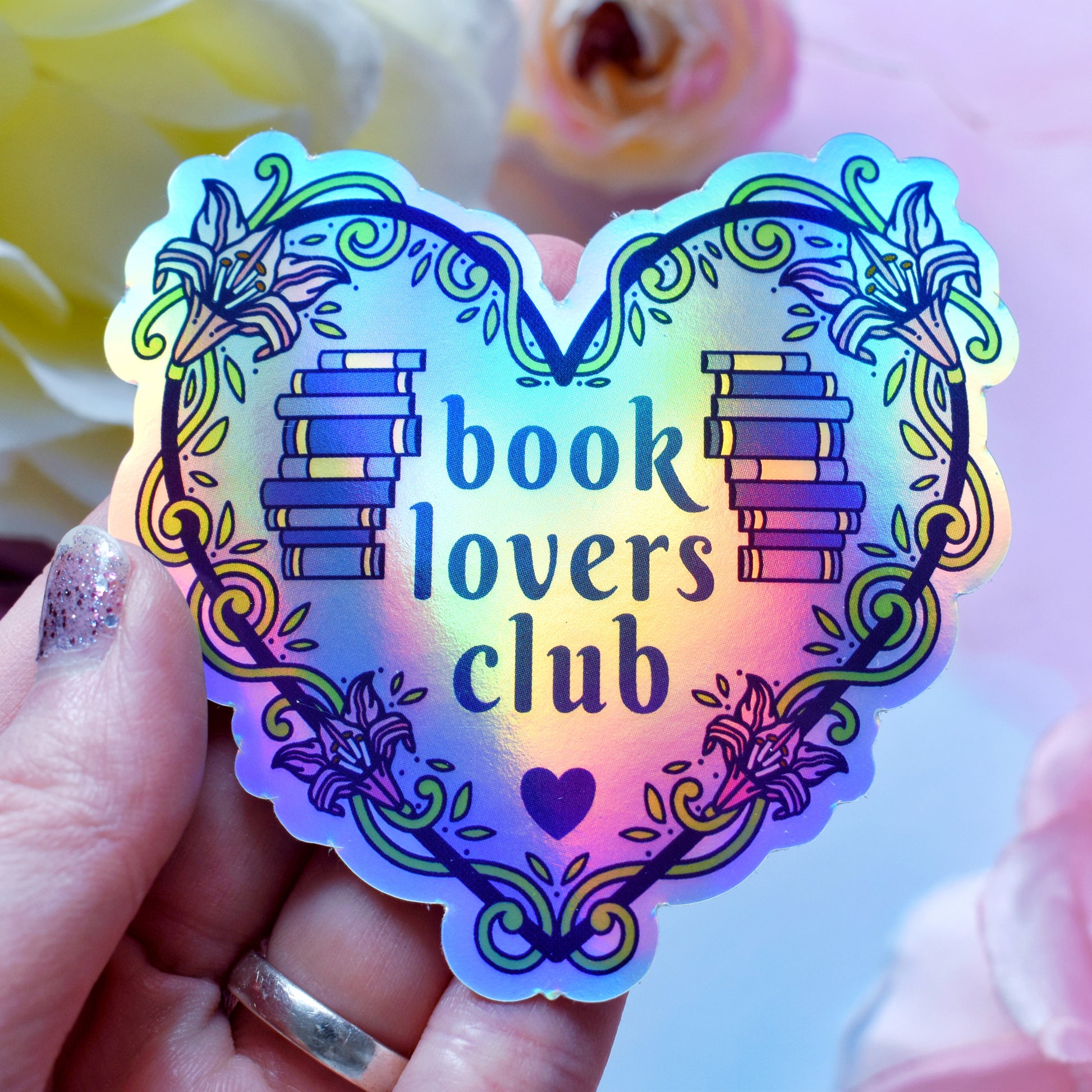 Blue heart-shaped bookish holographic sticker design features text 'book lovers club' with a floral and swirly outline with book spine stack. Held by a white hand above a watercolour style background in blue, pink and purples with flowers around the edge of the photo.