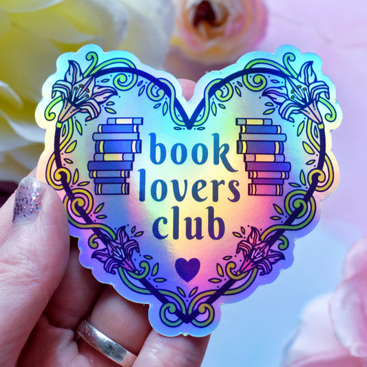 Blue heart-shaped bookish holographic sticker design features text 'book lovers club' with a floral and swirly outline with book spine stack. Held by a white hand above a watercolour style background in blue, pink and purples with flowers around the edge of the photo.