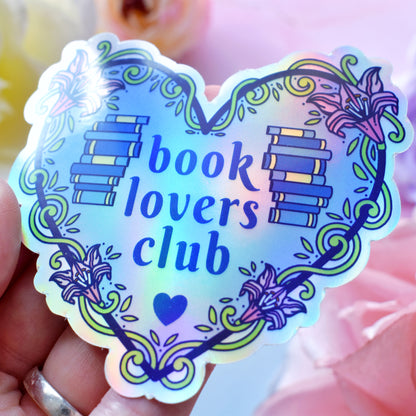 Blue heart-shaped bookish holographic sticker design features text 'book lovers club' with a floral and swirly outline with book spine stack. Held at an angle by a white hand above a watercolour style background in blue, pink and purples with flowers around the edge of the photo.