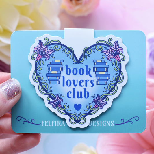 Blue heart-shaped design with a white border, featuring text 'book lovers club' with a floral and swirly outline with book spine stacks folded over a teal card. Card held by a white hand above a watercolour style background in blue, pink and purples with flowers around the edge of the photo.