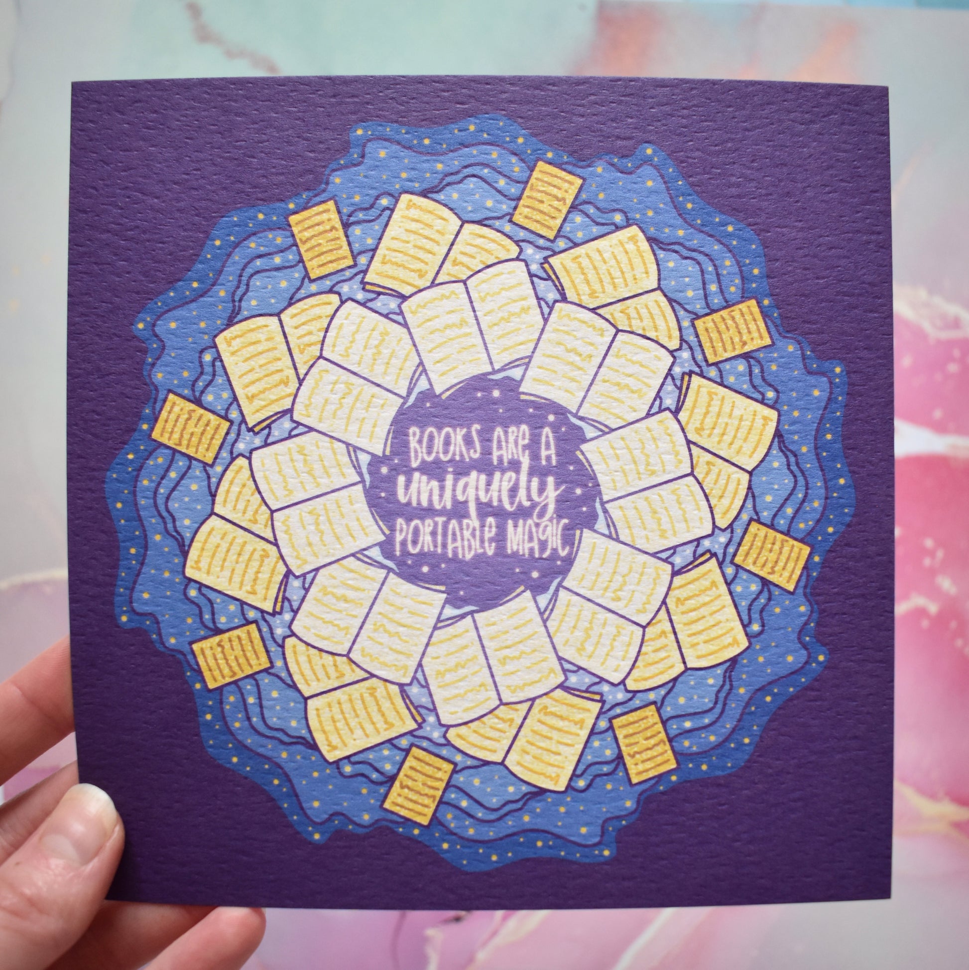 White hand holding print. Square print featuring a repeating pattern of yellow open books and blue swirls with purple around the edge. Design reads books are a uniquely portable magic in yellow at the centre of the design. Background of photograph is a pastel pink/blue watercolour style.