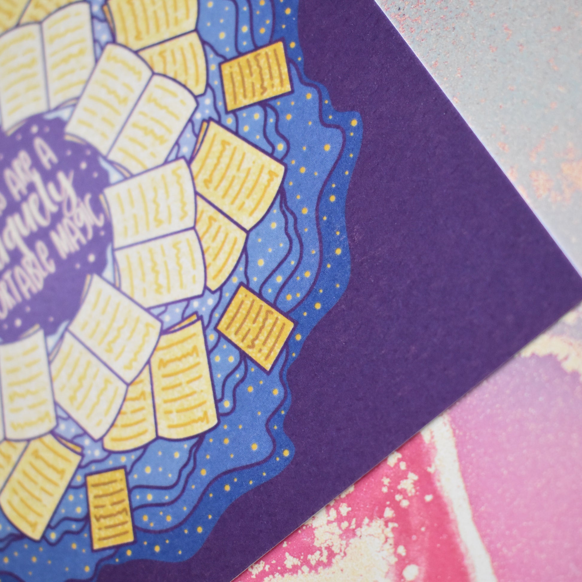 Close up of corner of design to show the texture of the print which is slightly raised. Square print featuring a repeating pattern of yellow open books and blue swirls with purple around the edge. Design reads books are a uniquely portable magic in yellow at the centre of the design. Background of photograph is a pastel pink/blue watercolour style.
