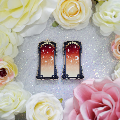 2 wodden charms, 1 with a loop at the top, 1 without next to each other on a sparkly background surrounded by fake flowers. Designs have black detailed frame. Gradient colour yellow through to red with stars and a moon inside frame.