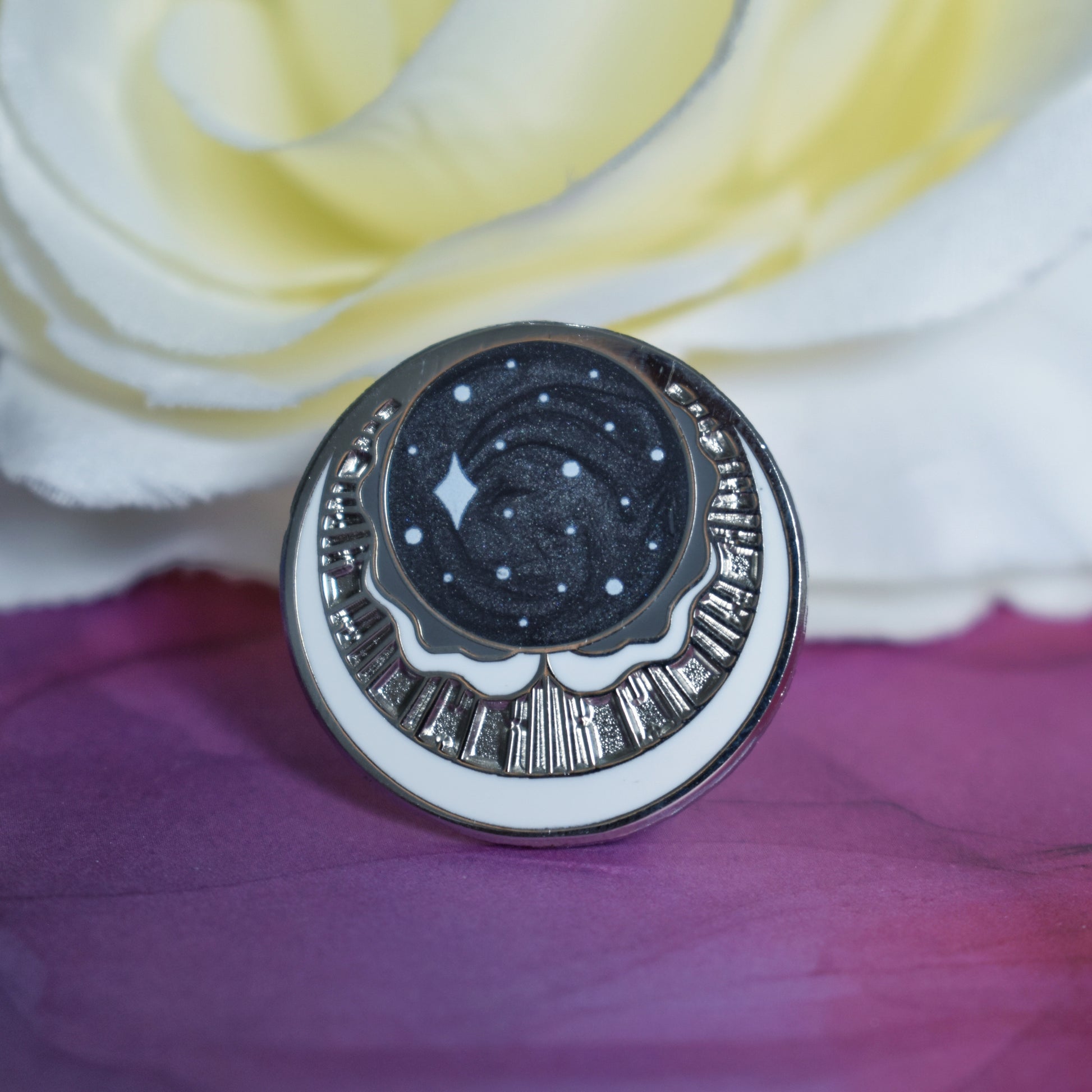 Silver metal enamel pin in centre of image. Enamel pin has a swirly black pearl circle top centre with some sparkles on. under is a cresent moon shape with raised metal liners and textured recessed metal areas. Under that and up the sides is a white cresent moon. Background of photograph is dark pink/purple water inspired with a fake cream flower.