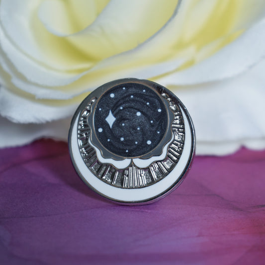 Silver metal enamel pin in centre of image. Enamel pin has a swirly black pearl circle top centre with some sparkles on. under is a cresent moon shape with raised metal liners and textured recessed metal areas. Under that and up the sides is a white cresent moon. Background of photograph is dark pink/purple water inspired with a fake cream flower.
