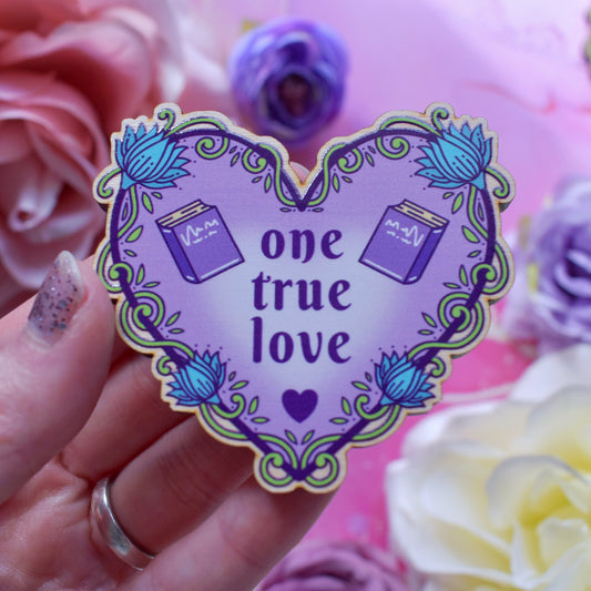 Purple heart-shaped bookish pin/magnet version, design features text 'one true love' with a floral and swirly outline with small front facing books. Pin/magnet held by a white hand above a watercolour style background in peachy pinks with flowers around the edge of the photo.
