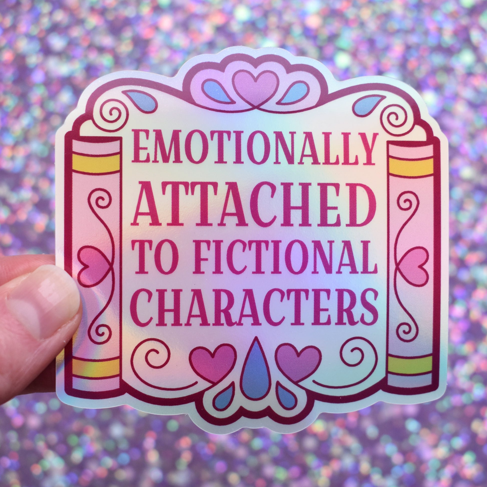 Holographic bookish sticker with text 'Emotionally Attached to Fictional Characters' designed to look somewhat like a wedding arch but made up of books either side and lots of hearts and teardrops. Held by a white hand against a glittery purple background.