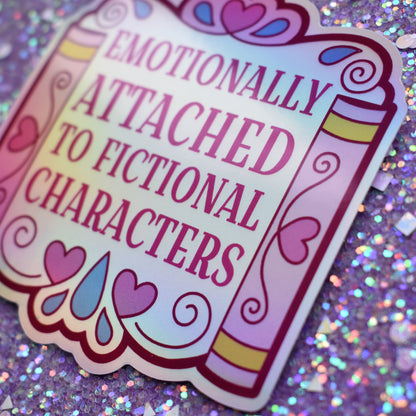 Close up of holographic bookish sticker with text 'Emotionally Attached to Fictional Characters' designed to look somewhat like a wedding arch but made up of books either side and lots of hearts and teardrops. Held by a white hand against a glittery purple background