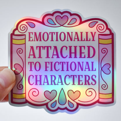 Holographic bookish sticker with text 'Emotionally Attached to Fictional Characters' designed to look somewhat like a wedding arch but made up of books either side and lots of hearts and teardrops. Held by a white hand against a an almost white background.