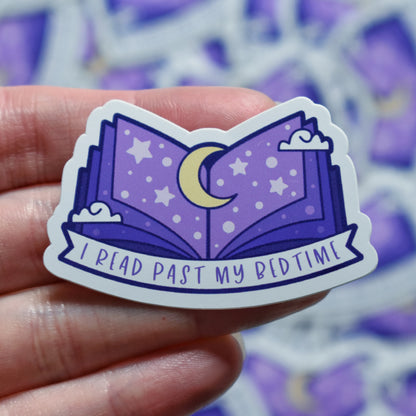 White hand holding sticker. Sticker design is an open book with a purple gradient, with a yellow moon top middle and white dots/stars/clouds spread across the design. A white banner at the bottom reads I read past my bedtime. Sticker has a white border. Background is blurry/distance of same stickers.